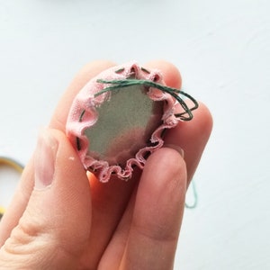 Puede incluir: Un c&iacute;rculo de tela rosa con un borde rizado, cosido alrededor de un c&iacute;rculo gris. La tela est&aacute; cosida con hilo verde.