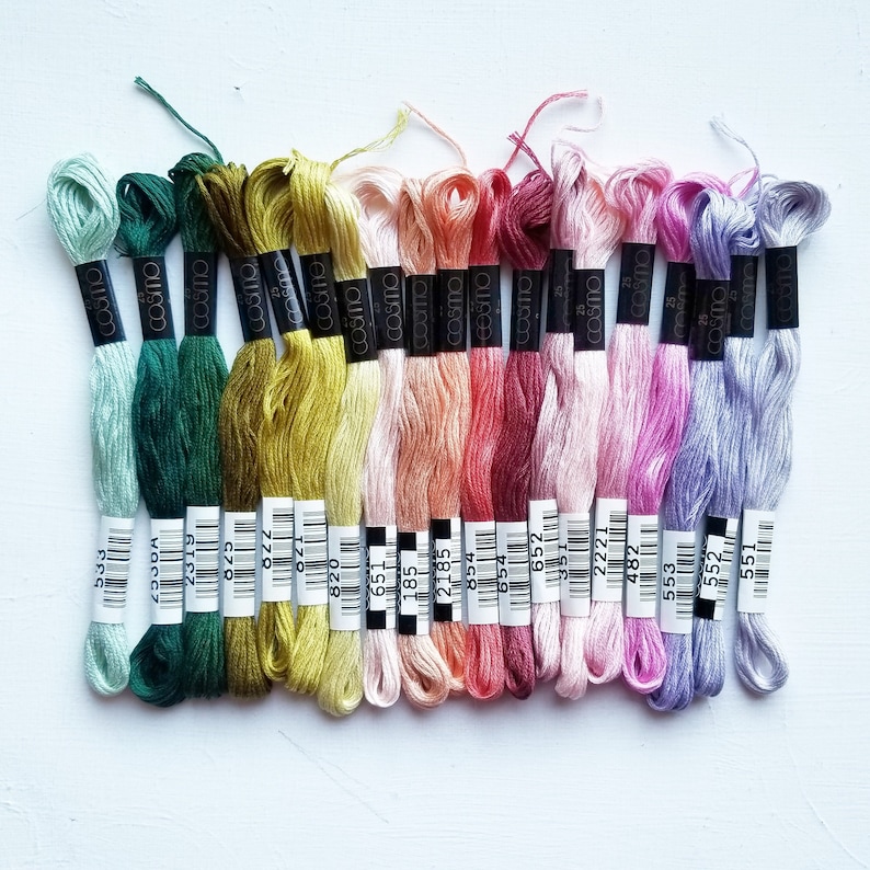 Pode incluir: Uma cole&ccedil;&atilde;o de 18 novelos de fio para bordar em v&aacute;rios tons de verde, amarelo, rosa, vermelho e roxo. O fio est&aacute; disposto em uma fila, com as etiquetas voltadas para o observador. As etiquetas s&atilde;o pretas com texto branco e incluem o nome da marca "cosmo" e um n&uacute;mero.