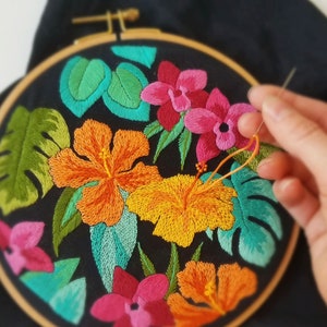 May include: A close-up of a colourful embroidery hoop with a floral design. The design features pink, orange, and green flowers on a black background.