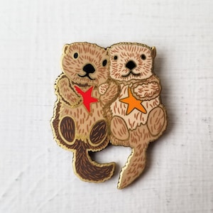 May include: Two brown and tan sea otters holding a red and orange star. The otters are holding each other and smiling. The otters are made of metal and have a shiny finish.