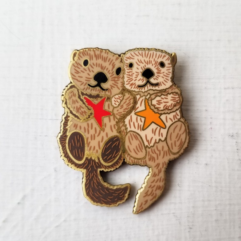 May include: Two brown and tan sea otters holding a red and orange star. The otters are holding each other and smiling. The otters are made of metal and have a shiny finish.