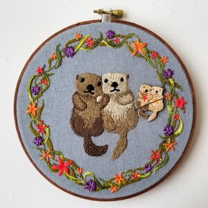 May include: Embroidered hoop art featuring two brown and white otters holding paws. The otters are surrounded by a wreath of green leaves and orange flowers. Two smaller otters are embroidered below the larger otters.