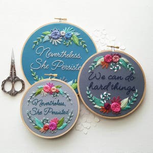 Może przedstawiać: Trzy haftowane obręcze z inspirującymi cytatami. Największa obręcz jest niebieska z cytatem "Nevertheless, She Persisted". Środkowa obręcz jest szara z cytatem "We can do hard things". Najmniejsza obręcz jest szara z cytatem "Nevertheless, She Persisted".