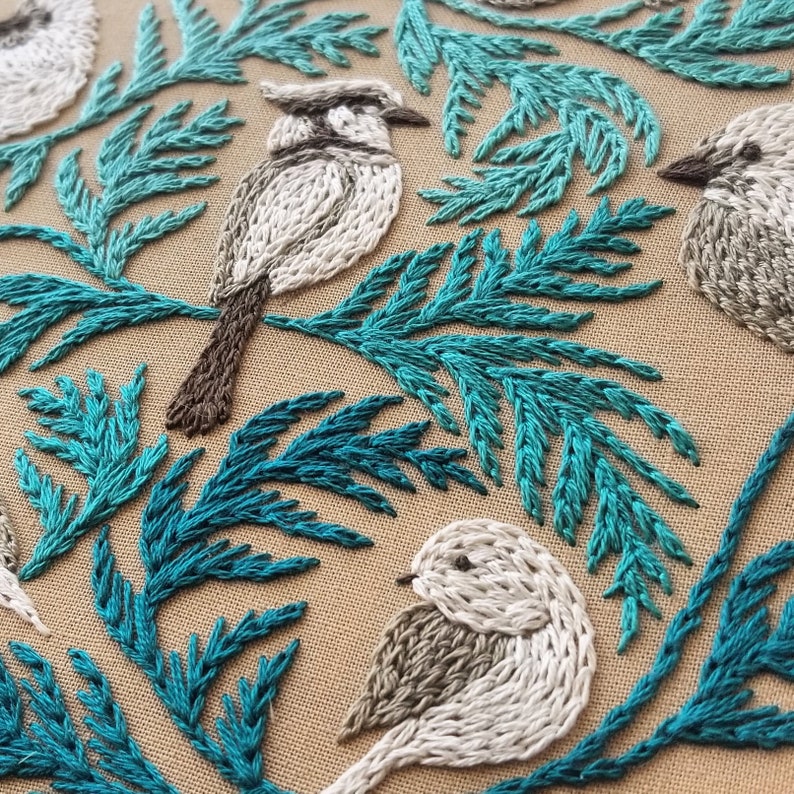 以下が含まれることがあります： 白鳥と茶色のくちばし、足が特徴の刺繍作品。ターコイズグリーンの葉っぱに囲まれています。鳥は、1本1本の羽根がわかるほど、リアルな刺繍で表現されています。