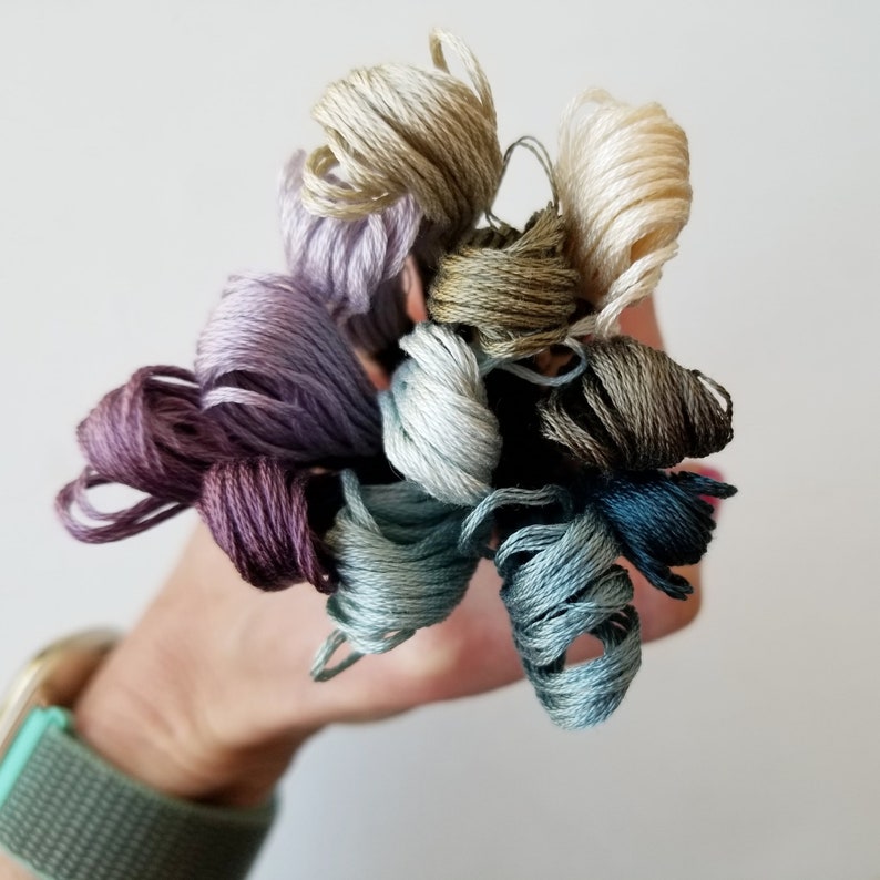 May include: A collection of embroidery floss spools in various shades of purple, blue, green, and brown. The floss is wound into small balls and held in a hand.