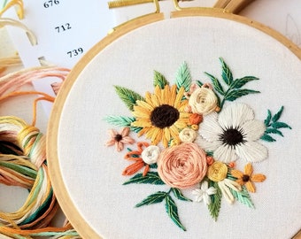 May include: A floral embroidery hoop featuring a sunflower, white anemone, and other flowers in shades of yellow, orange, and peach. The embroidery is set in a wooden hoop. A card with numbers 564, 676, 712, and 739 is visible, along with colorful embroidery floss.