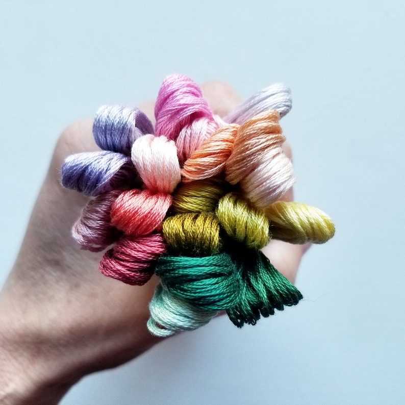 Pode incluir: Uma cole&ccedil;&atilde;o de carret&eacute;is de fio de bordado em v&aacute;rias cores, incluindo rosa, roxo, verde, amarelo e laranja. Os carret&eacute;is est&atilde;o dispostos em um padr&atilde;o circular e s&atilde;o mantidos juntos por uma m&atilde;o.