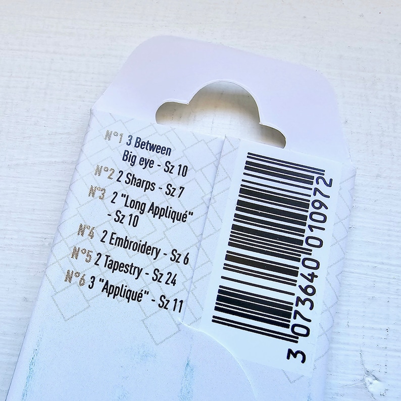 May include: Close-up of a white product label with black text listing different needle sizes and types. The label also includes a black and white barcode with the number 3073640 010972.