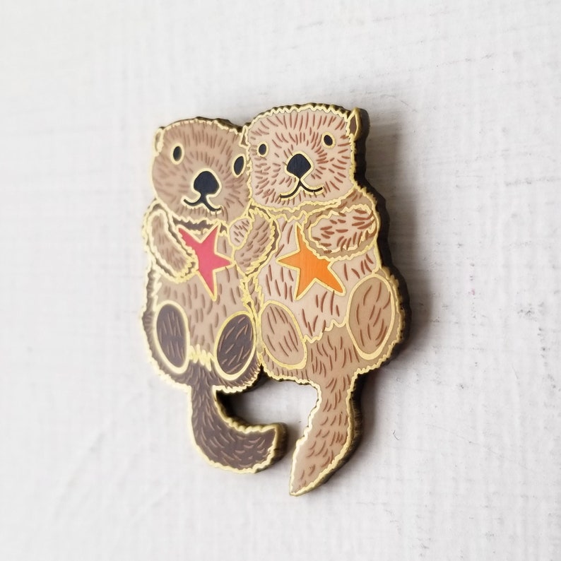 May include: A gold-tone enamel pin featuring two otters holding red and orange stars. The otters are brown and have black eyes and noses.