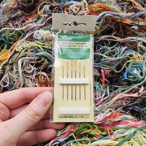 May include: A package of Clover Gold Eye Tapestry Needles, sizes 18, 20, and 22. The needles are gold-colored with a large eye, displayed on a white card. The background is a colorful assortment of embroidery floss.