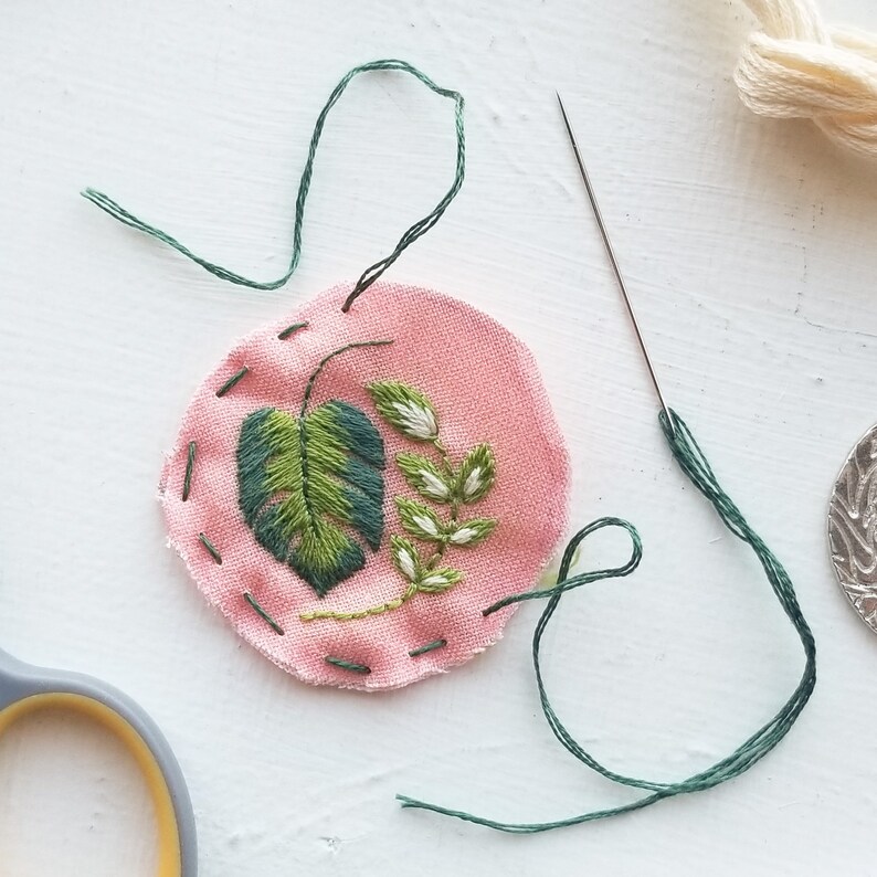 Puede incluir: Un c&iacute;rculo de tela rosa con bordado verde de una hoja y un tallo. La tela est&aacute; cosida alrededor del borde con hilo verde. Tambi&eacute;n se ve una aguja e hilo verde.
