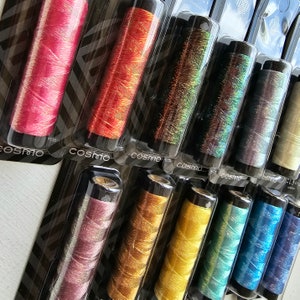 May include: A collection of Cosmo brand embroidery floss in various colors, including pink, orange, green, and blue. Each spool is packaged in a clear plastic container. The floss has a metallic, shimmering appearance.