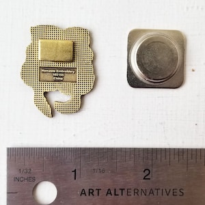 May include: Gold-colored metal pin with a floral design. The back of the pin has a magnetic backing and text that reads "Namaste Embroidery 102155 China".