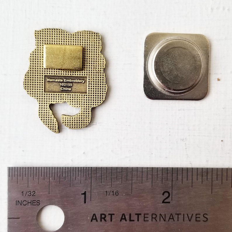 May include: Gold-colored metal pin with a floral design. The back of the pin has a magnetic backing and text that reads "Namaste Embroidery 102155 China".