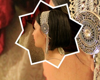 May include: A silver and white hair accessory with a decorative round pendant. The accessory is made of lace and features a silver metal design.