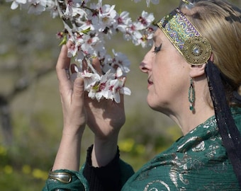 May include: A woman wearing a green and gold patterned shawl and a headband with a gold and blue design, is smelling a branch of white almond blossoms.