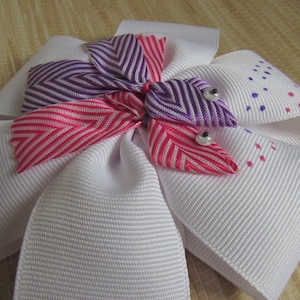 Puede incluir: Un lazo blanco con acentos de cinta a rayas rosa, morado y blanco. El lazo tiene dos adornos blancos con forma de perla.
