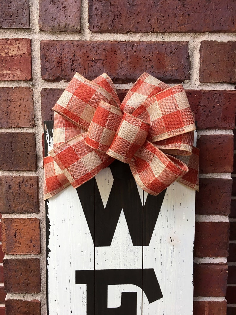 May include: A white wooden sign with the word "WELCOME" painted in black. A large red and white gingham bow is tied to the top of the sign.