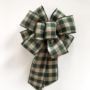 May include: A large decorative bow made from green and tan plaid ribbon. The bow has multiple loops and long tails, with a black stitched edge. The bow is centered on a white background.