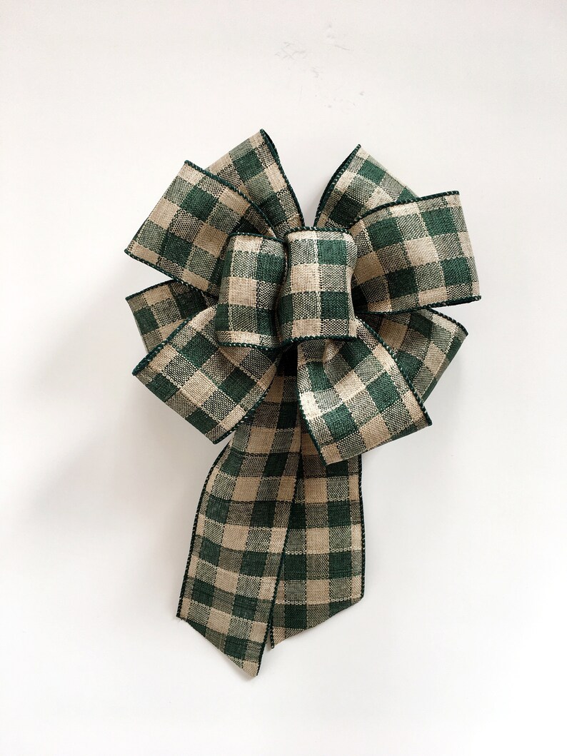 May include: A large decorative bow made from green and tan plaid ribbon. The bow has multiple loops and long tails, with a black stitched edge. The bow is centered on a white background.