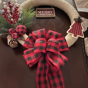 May include: A Christmas wreath made of natural twine with a red and black buffalo plaid bow, a small pine sprig, red berries, and a small buffalo plaid Christmas tree ornament. The wreath also has a sign that reads "Merry Christmas".