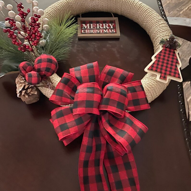 May include: A Christmas wreath made of natural twine with a red and black buffalo plaid bow, a small pine sprig, red berries, and a small buffalo plaid Christmas tree ornament. The wreath also has a sign that reads "Merry Christmas".