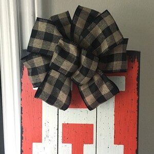 May include: A decorative wooden sign with a distressed red background and large white letters. A large bow made of black and tan plaid ribbon is attached to the top of the sign. The sign is likely for home decor.