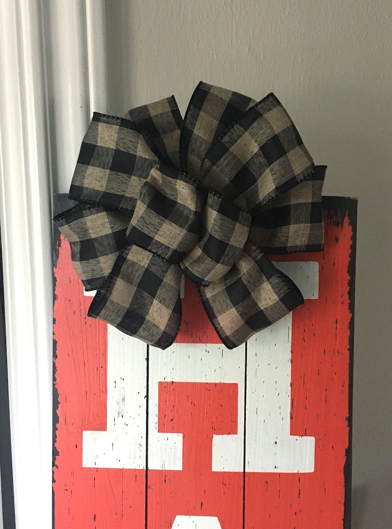 May include: A decorative wooden sign with a distressed red background and large white letters. A large bow made of black and tan plaid ribbon is attached to the top of the sign. The sign is likely for home decor.