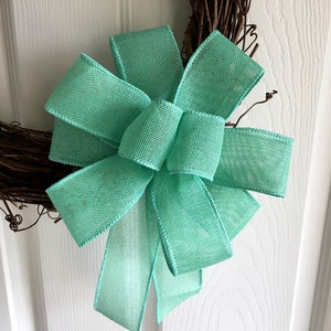 May include: A large, light green burlap bow tied on a grapevine wreath.