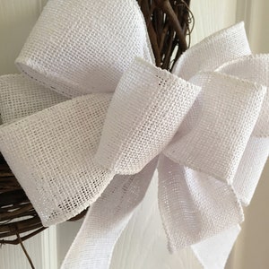 May include: A white burlap bow with a woven texture is tied around a brown twig wreath.