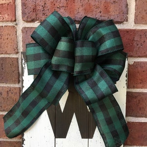 May include: A wooden sign with the word "Welcome" painted in black on a white background. A large green and black plaid bow is tied to the top of the sign.