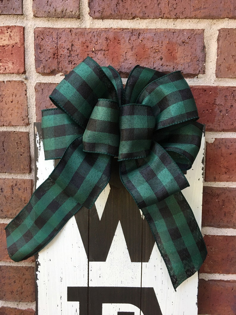 May include: A wooden sign with the word "Welcome" painted in black on a white background. A large green and black plaid bow is tied to the top of the sign.