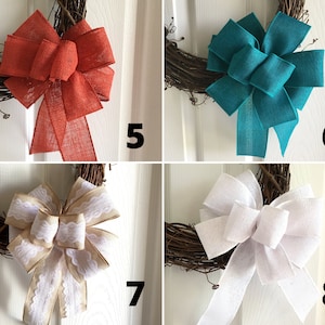 May include: Four different burlap bows in various colors, including red, teal, beige, and white, are shown on a wreath. The bows are tied with a simple knot and have a rustic, farmhouse style.