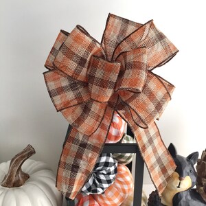 May include: A large decorative bow made of plaid ribbon in shades of orange, brown, and beige. The bow sits atop a black lantern filled with decorative pumpkins in various plaid patterns. A white pumpkin and a squirrel figurine are also visible.