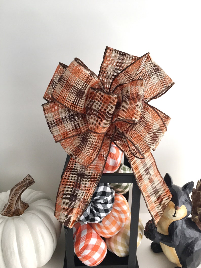 May include: A large decorative bow made of plaid ribbon in shades of orange, brown, and beige. The bow sits atop a black lantern filled with decorative pumpkins in various plaid patterns. A white pumpkin and a squirrel figurine are also visible.