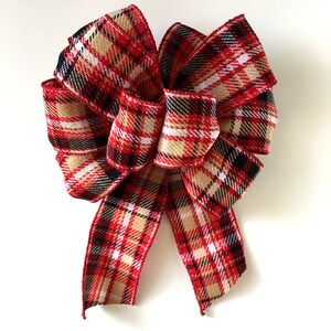 May include: A decorative bow with a classic plaid pattern in red, black, white, and tan. The bow is made of a textured fabric and is suitable for holiday or seasonal decor. The bow is tied with multiple loops and tails.