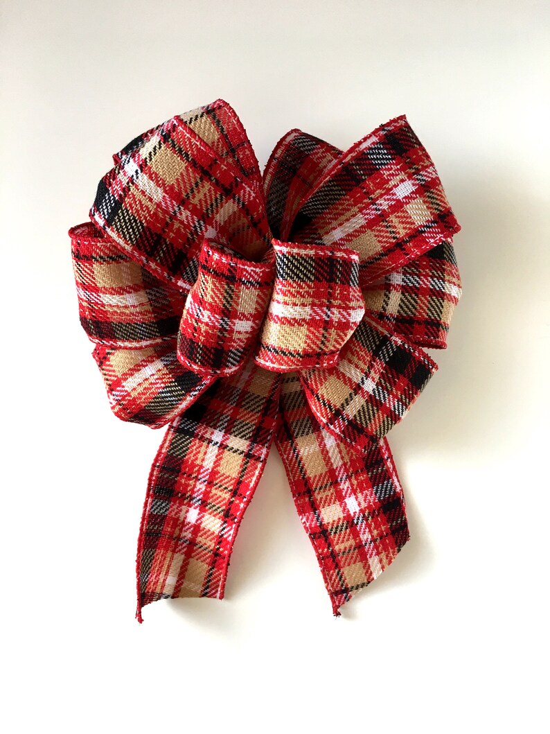 May include: A decorative bow with a classic plaid pattern in red, black, white, and tan. The bow is made of a textured fabric and is suitable for holiday or seasonal decor. The bow is tied with multiple loops and tails.