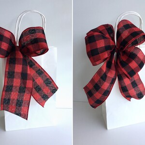 May include: A white gift bag with a large red and black buffalo plaid bow. The bow is made of a sheer ribbon and is tied at the top of the bag. The bag has white handles.