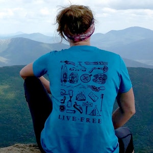 May include: A person wearing a blue t-shirt with a graphic of camping and outdoor icons and the text "LIVE-FREE". The person is sitting on a rocky outcrop overlooking a mountain range.
