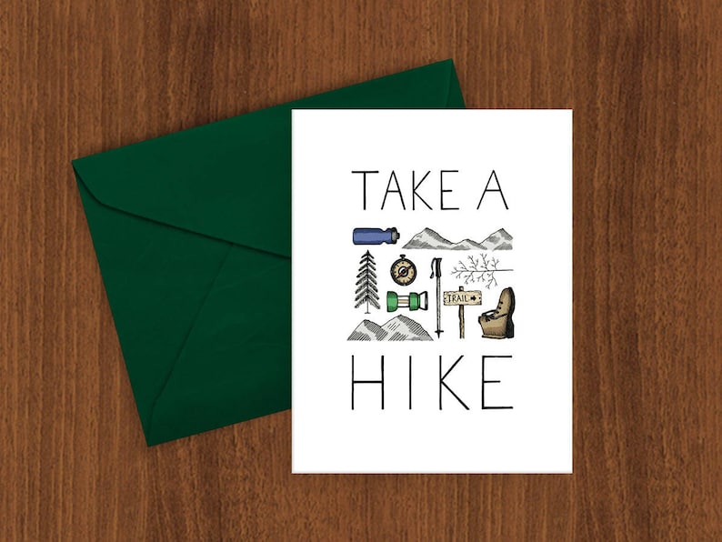 May include: A white card with the words "TAKE A HIKE" and hiking-themed illustrations, including a water bottle, compass, mountains, and a trail sign. A dark green envelope is on the left side of the card. The card is on a wooden surface.