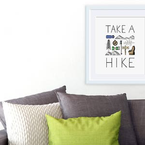 May include: A framed print with the words "TAKE A HIKE" in black lettering. The print features hiking-themed illustrations in blue, green, and brown. The frame is white, and the print is displayed above a grey sofa with cushions.
