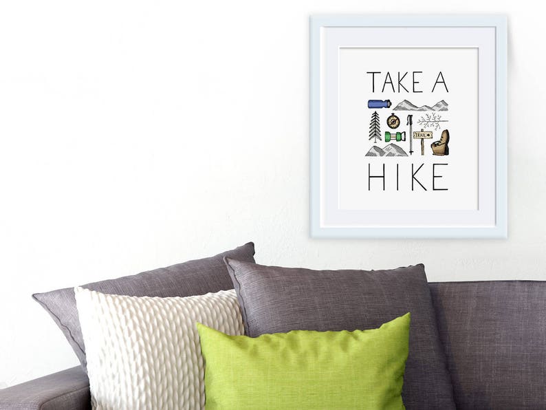 May include: A framed print with the words "TAKE A HIKE" in black lettering. The print features hiking-themed illustrations in blue, green, and brown. The frame is white, and the print is displayed above a grey sofa with cushions.