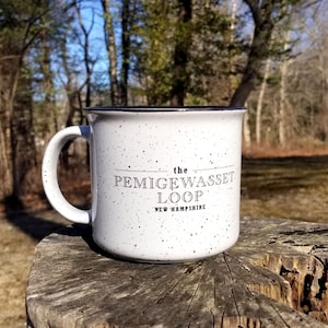 May include: White camping mug with black speckled glaze and black text that reads "the Pemigewasset Loop New Hampshire". The mug is sitting on a tree stump in a forest setting.