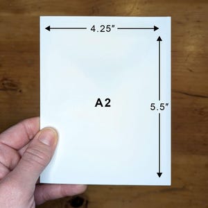 May include: A white A2 card, measuring 10.8 cm wide and 14 cm tall, is held by a hand. The card has the text "A2" printed on it. The background is a wooden surface.