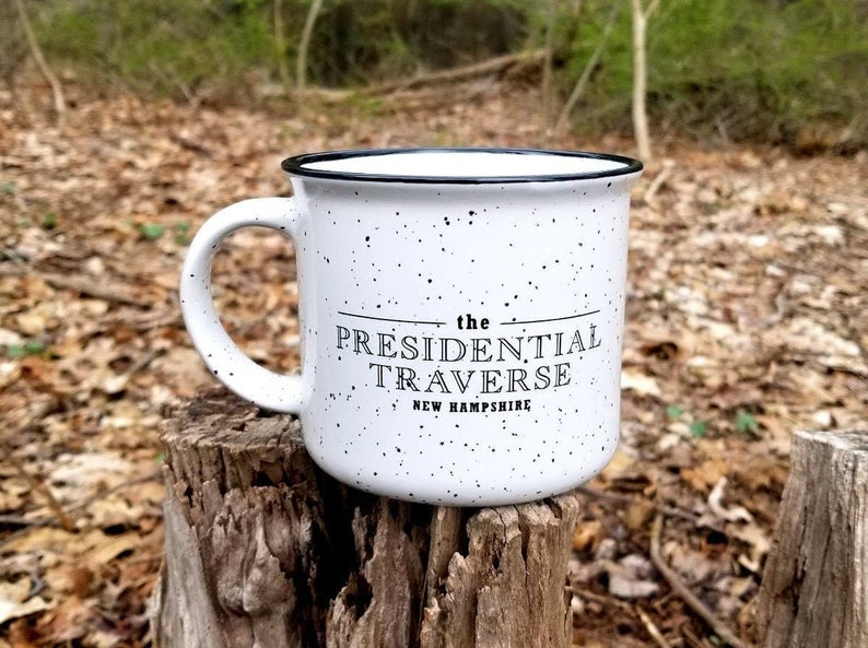Puede incluir: Taza de cer&aacute;mica blanca moteada con borde y asa negros. La taza presenta el texto "the PRESIDENTIAL TRAVERSE NEW HAMPSHIRE" en letras negras. La taza est&aacute; sobre un toc&oacute;n de &aacute;rbol al aire libre.
