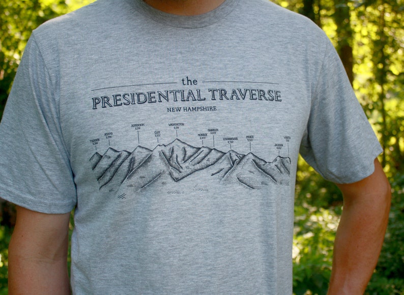 Op de afbeelding: Een grijs T-shirt met een zwarte afbeelding van de Presidential Traverse in New Hampshire. De afbeelding toont de bergketen met de namen van de toppen en hun hoogtes.