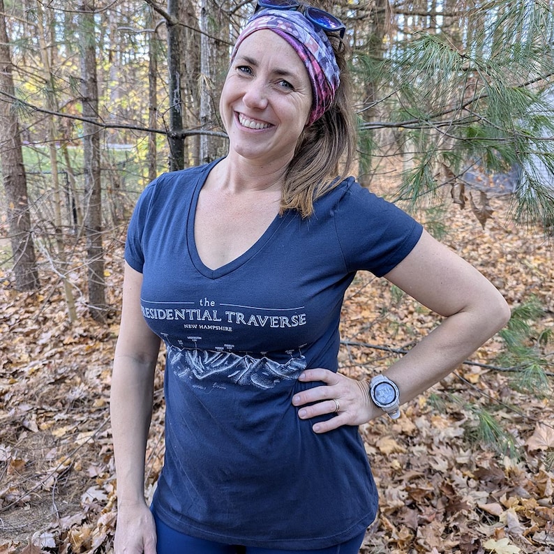 Op de afbeelding: Een vrouw draagt een donkerblauw V-hals T-shirt met een witte afbeelding van de Presidential Traverse in New Hampshire. Ze draagt een kleurrijk hoofdband en een zilveren horloge.