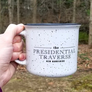 Puede incluir: Taza de cer&aacute;mica blanca moteada con borde y asa negros. La taza presenta el texto "the PRESIDENTIAL TRAVERSE NEW HAMPSHIRE" en letras negras. La taza se sostiene contra un fondo borroso al aire libre.