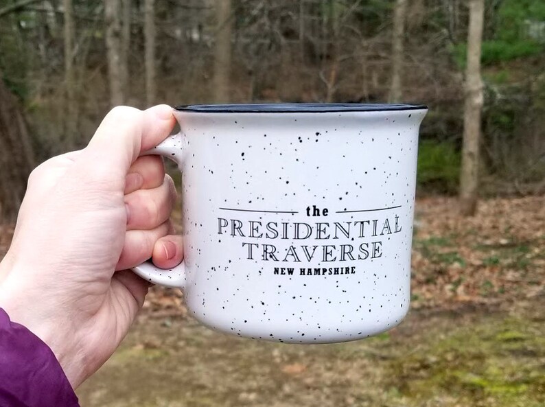 Puede incluir: Taza de cer&aacute;mica blanca moteada con borde y asa negros. La taza presenta el texto "the PRESIDENTIAL TRAVERSE NEW HAMPSHIRE" en letras negras. La taza se sostiene contra un fondo borroso al aire libre.
