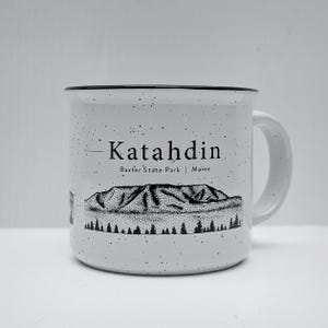 May include: White ceramic mug with a black speckled glaze. The mug features a black and white illustration of Katahdin Mountain in Baxter State Park, Maine.